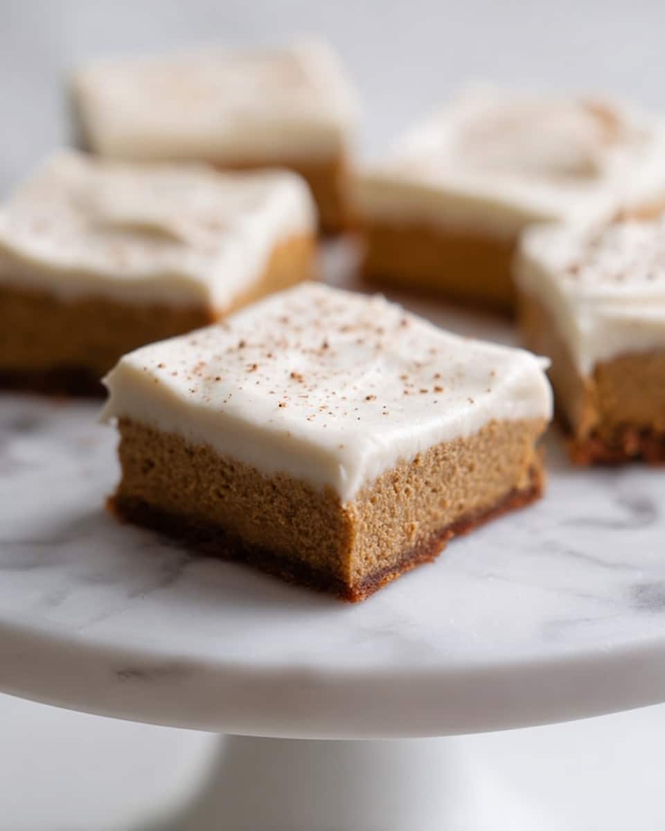 A close-up view of several square dessert bars on a white cake stand with a smooth finish, set against a white marbled background. Each bar has two layers: the bottom layer is thick, brown, and textured with a slightly crumbly surface, while the top layer is a thick, creamy white frosting with a smooth and soft texture. The frosting is sprinkled lightly with small brown specks evenly across the top. The bars are arranged with one in the foreground in clear focus and others blurred in the background. Photo taken with an iphone --ar 4:5 --v 7