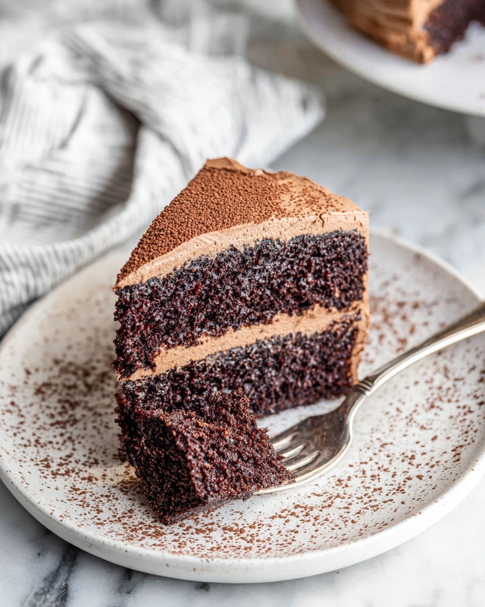 A slice of rich chocolate cake with two thick dark brown layers separated by smooth light brown frosting sits on a white plate speckled with cocoa powder. The outside of the cake is coated evenly with the same light brown frosting. A piece of the cake is cut and held by a silver fork placed at the edge of the plate. The plate rests on a white marbled surface, and a soft white and gray striped cloth is blurred in the background. Photo taken with an iphone --ar 4:5 --v 7