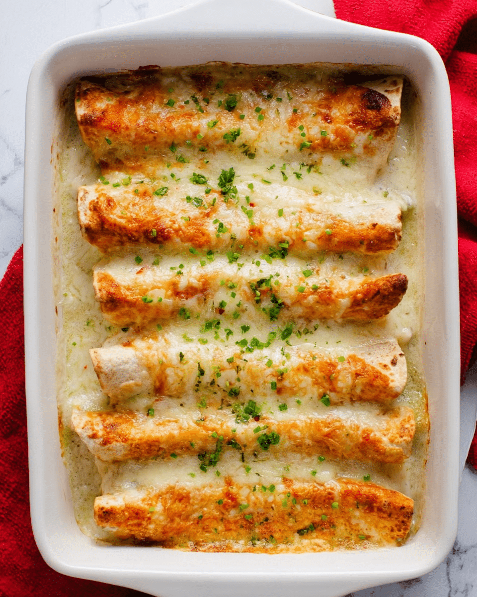The image shows a white square baking dish filled with five long rolled enchiladas covered in melted cheese. The enchiladas have a light brown, crispy layer on top with some green chopped herbs sprinkled over them. The cheese sauce underneath is creamy and pale yellow, covering the entire base of the dish. The baking dish is set on a white marbled surface with a red cloth partially visible on the left side. Photo taken with an iphone --ar 4:5 --v 7