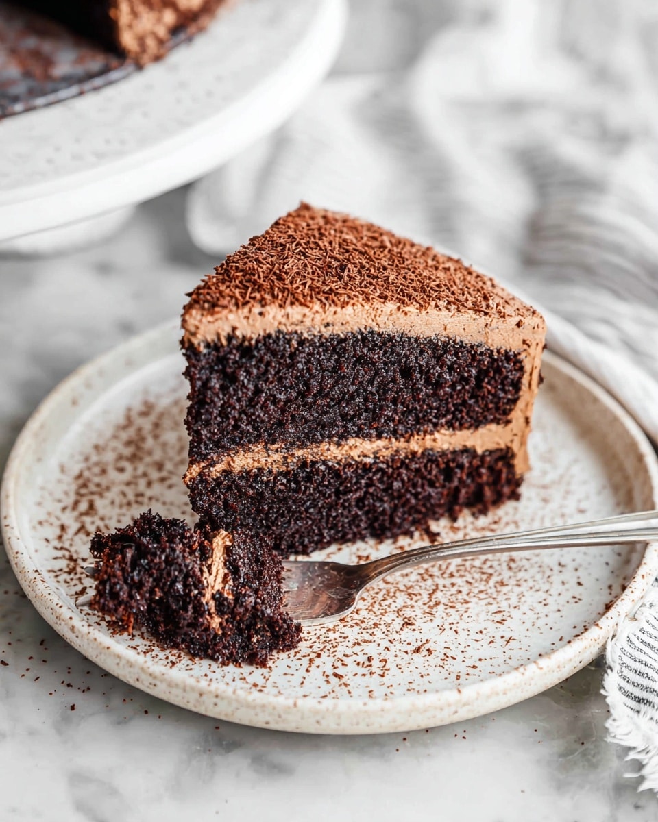 A slice of rich chocolate cake with two dark brown sponge layers separated by a thick layer of smooth, light brown chocolate frosting. The outer side of the cake slice is covered with the same light brown frosting, which looks creamy and soft. The cake is placed on a white plate with small brown cocoa powder sprinkles scattered around, adding texture to the scene. A silver fork has a small bite-sized piece of the cake on its tines, positioned in front of the slice. The background shows a white cloth with light gray stripes on a white marbled surface. photo taken with an iphone --ar 4:5 --v 7
